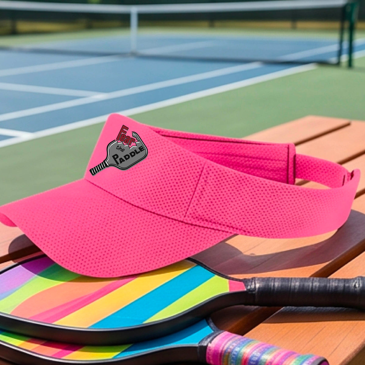 A close up of a pink visor with a 'Fear the Paddle' cutout from a pickleall paddle with a blurred pickleblal court in the background.