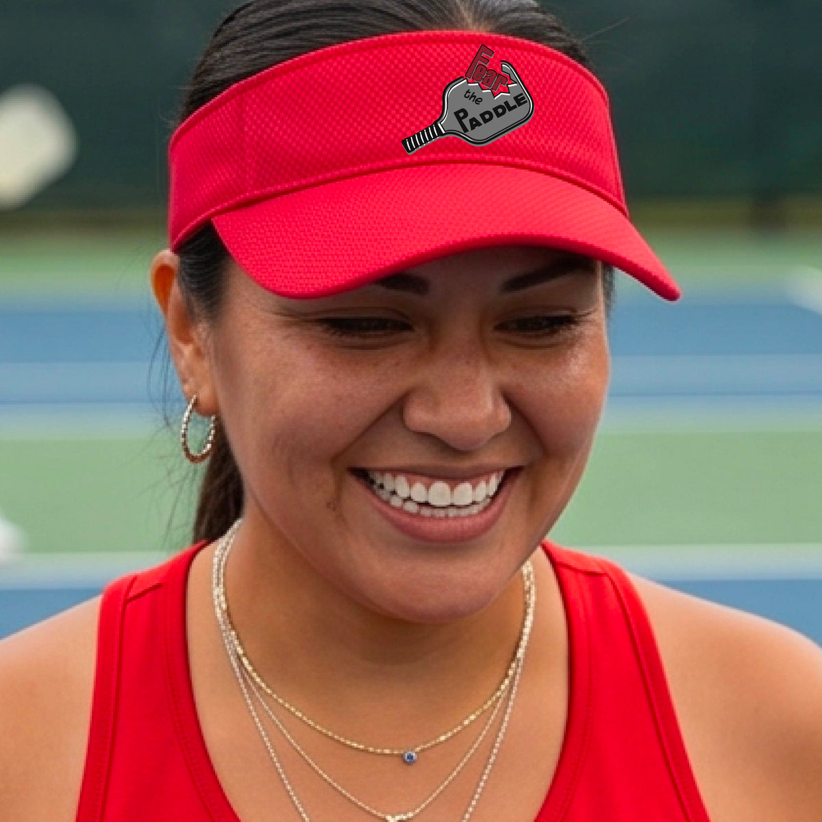 A close up of a bright red visor with a 'Fear the Paddle' cutout from a pickleall paddle with a blurred pickleblal court in the background.