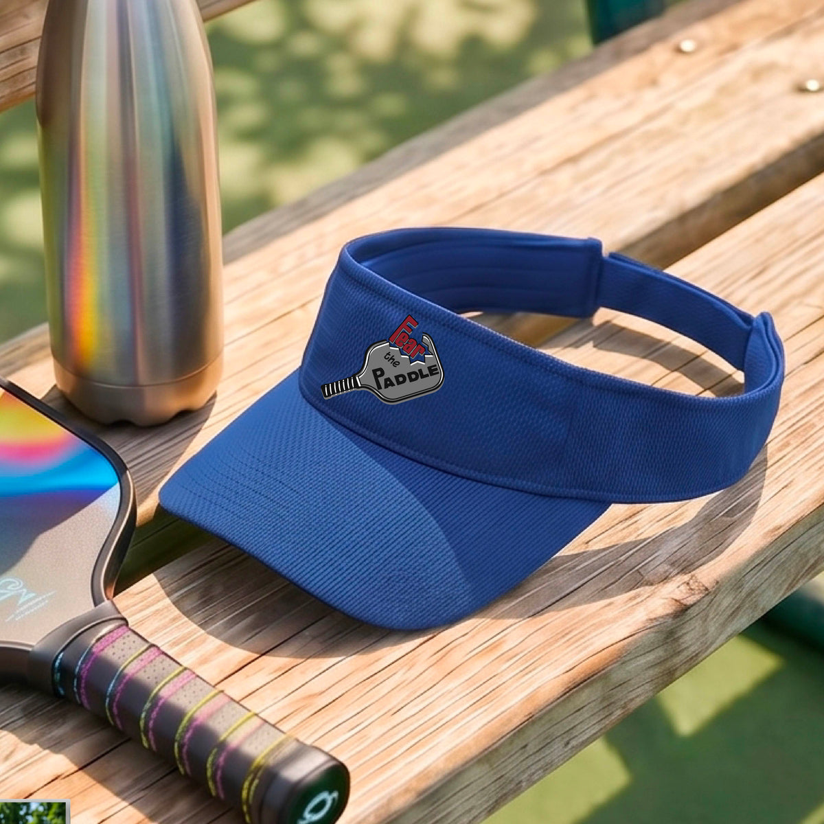 A close up of a royal blue visor with a 'Fear the Paddle' cutout from a pickleall paddle with a blurred pickleblal court in the background.
