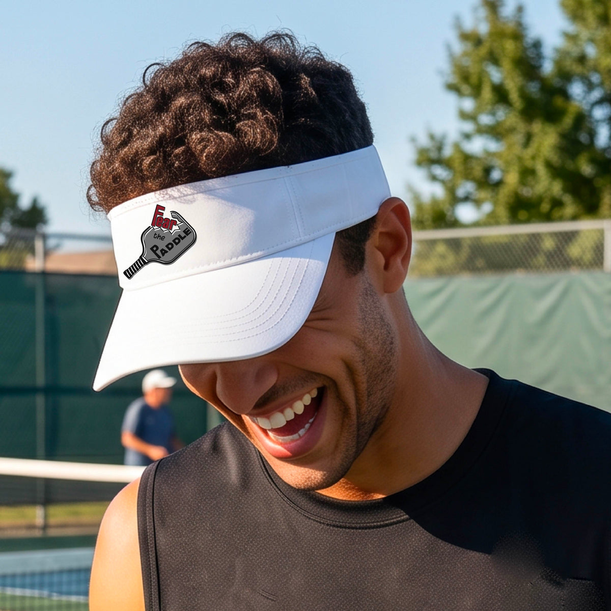 A close up of a white visor with a 'Fear the Paddle' cutout from a pickleall paddle with a blurred pickleblal court in the background.