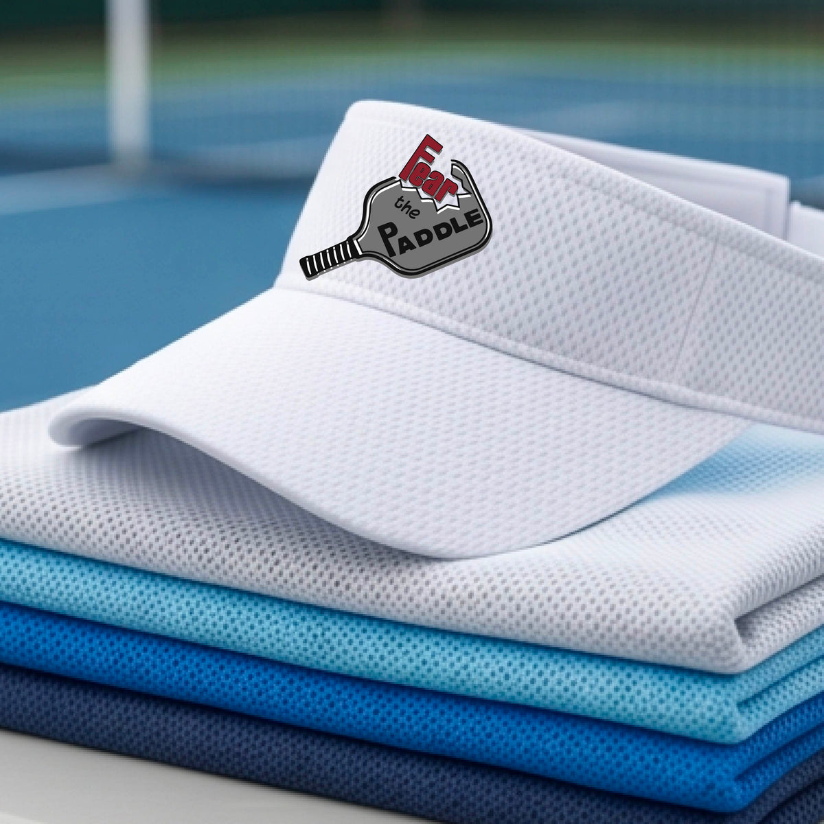 A close up of a white visor with a 'Fear the Paddle' cutout from a pickleall paddle with a blurred pickleblal court in the background.