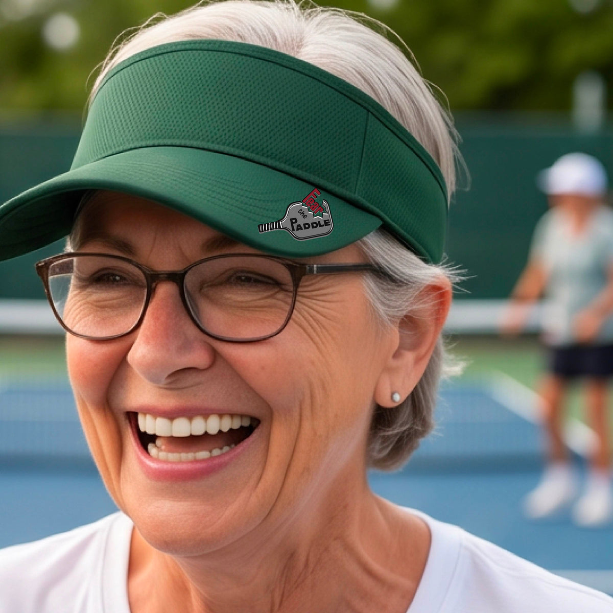 A close up of a green visor with a 'Fear the Paddle' cutout from a pickleall paddle with a blurred pickleblal court in the background.