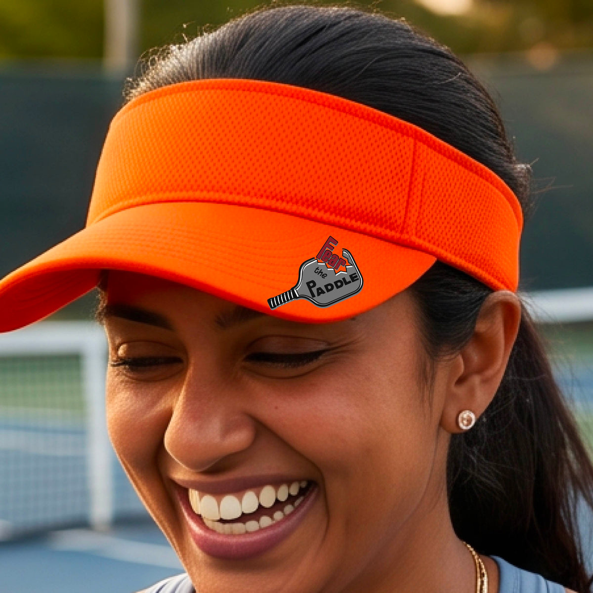 A close up of a bright orange visor with a 'Fear the Paddle' cutout from a pickleall paddle with a blurred pickleblal court in the background.