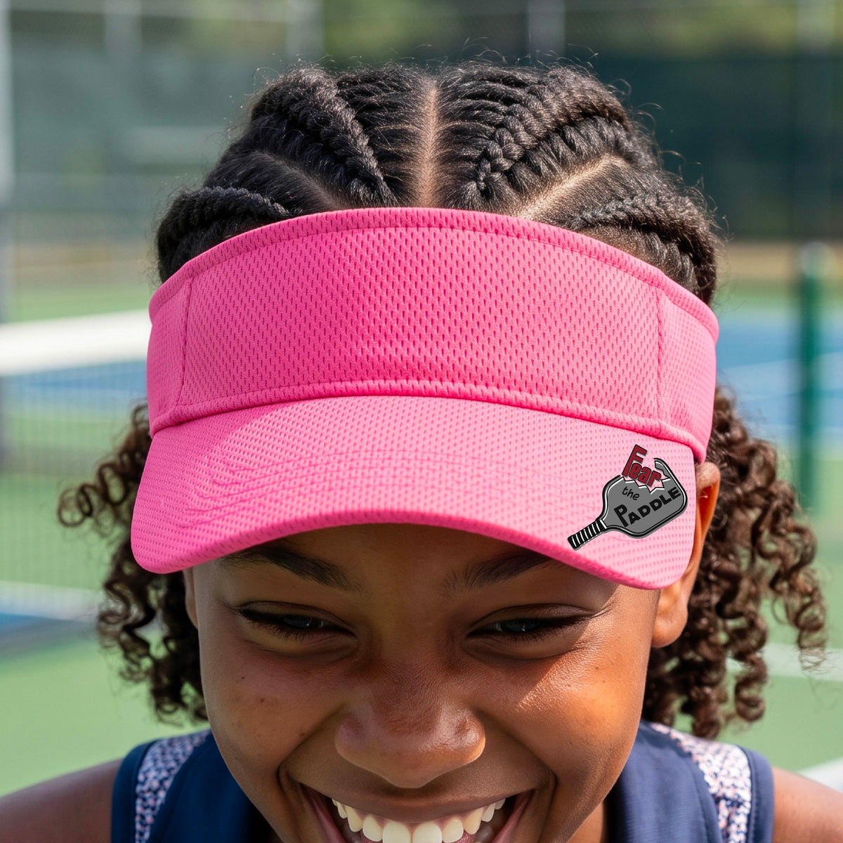 A close up of a pink visor with a 'Fear the Paddle' cutout from a pickleall paddle with a blurred pickleblal court in the background.