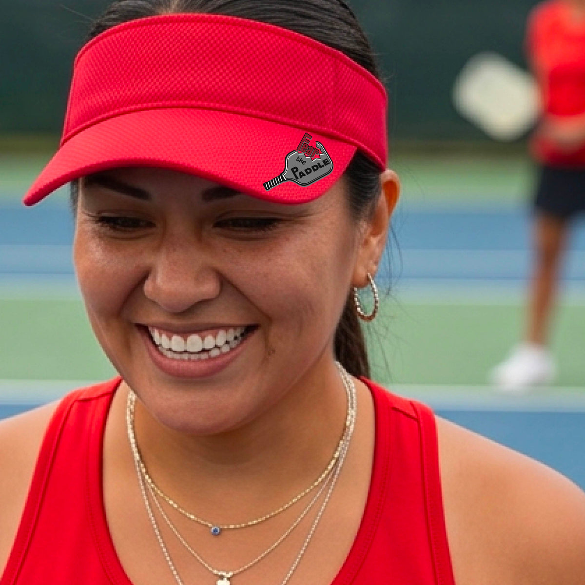 A close up of a bright red visor with a 'Fear the Paddle' cutout from a pickleall paddle with a blurred pickleblal court in the background.