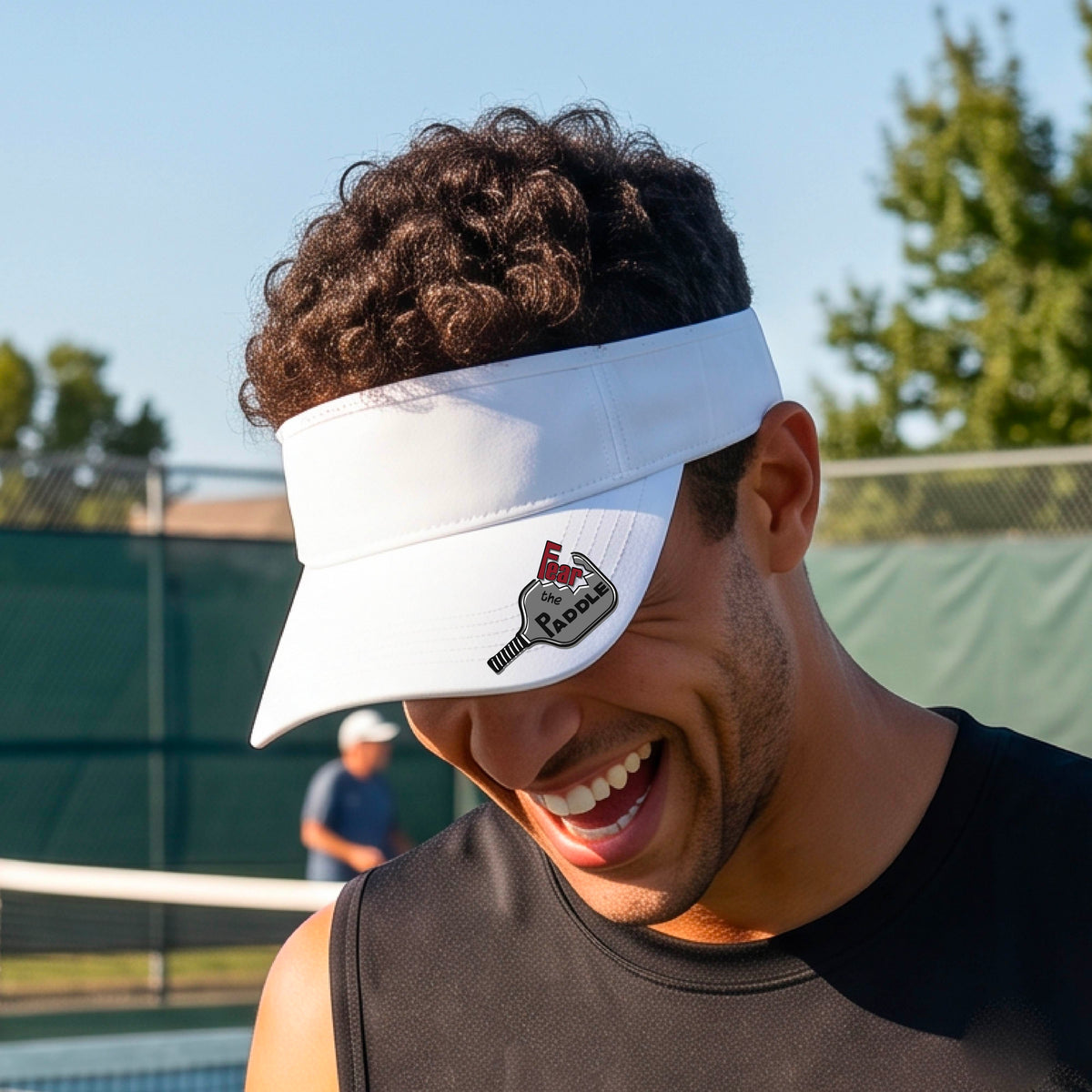 A close up of a white visor with a 'Fear the Paddle' cutout from a pickleall paddle with a blurred pickleblal court in the background.