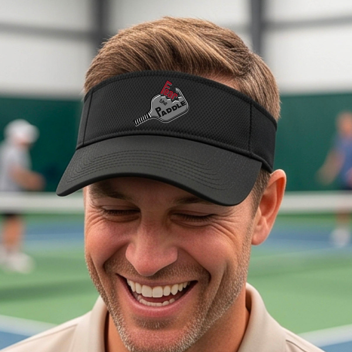 A close up of a black visor with a 'Fear the Paddle' cutout from a pickleall paddle with a blurred pickleblal court in the background.