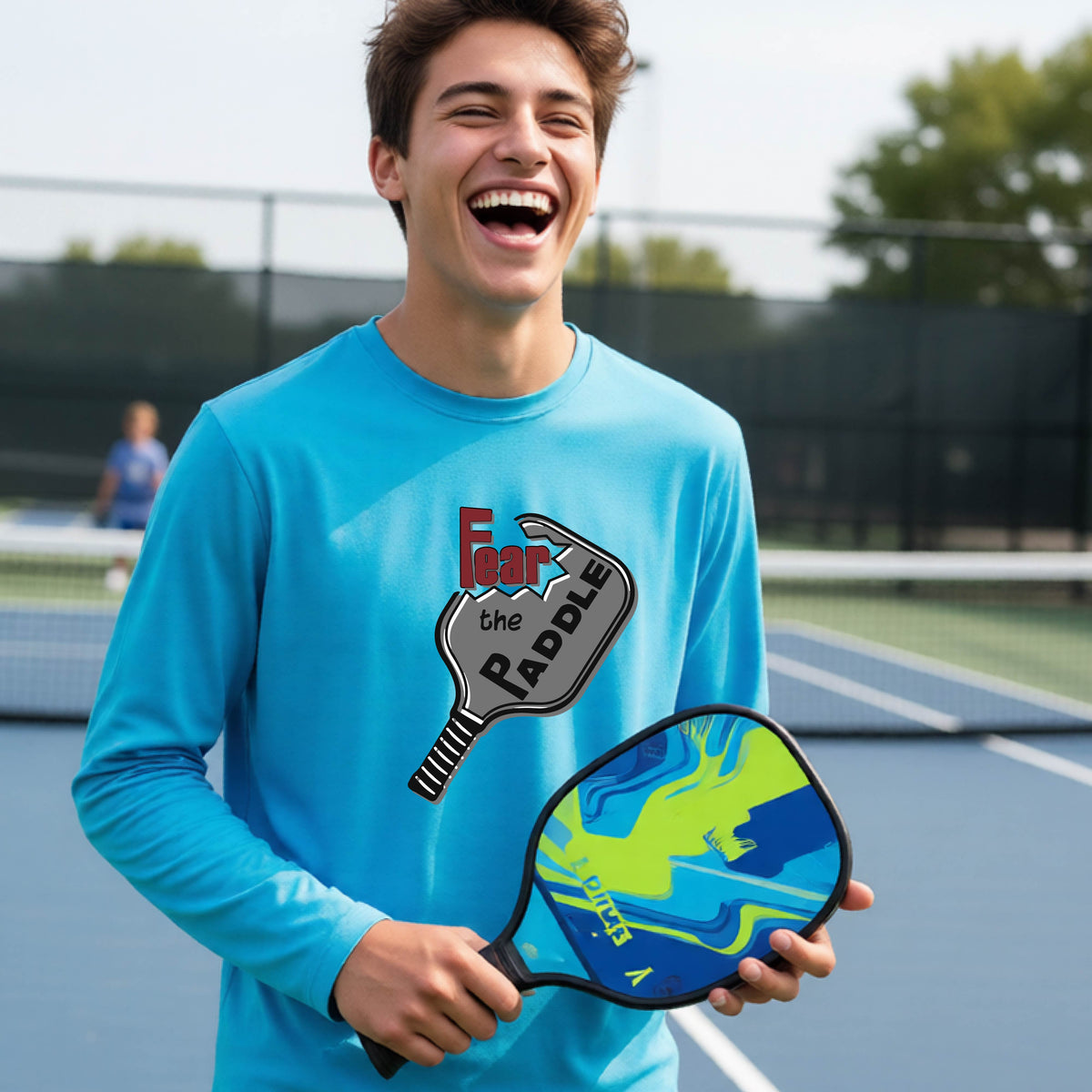 A young person wearing a light blue long-sleeve shirt with a pickleball graphic of the text 'FEAR THE PADDLE' cut out into a pickleball paddle. 