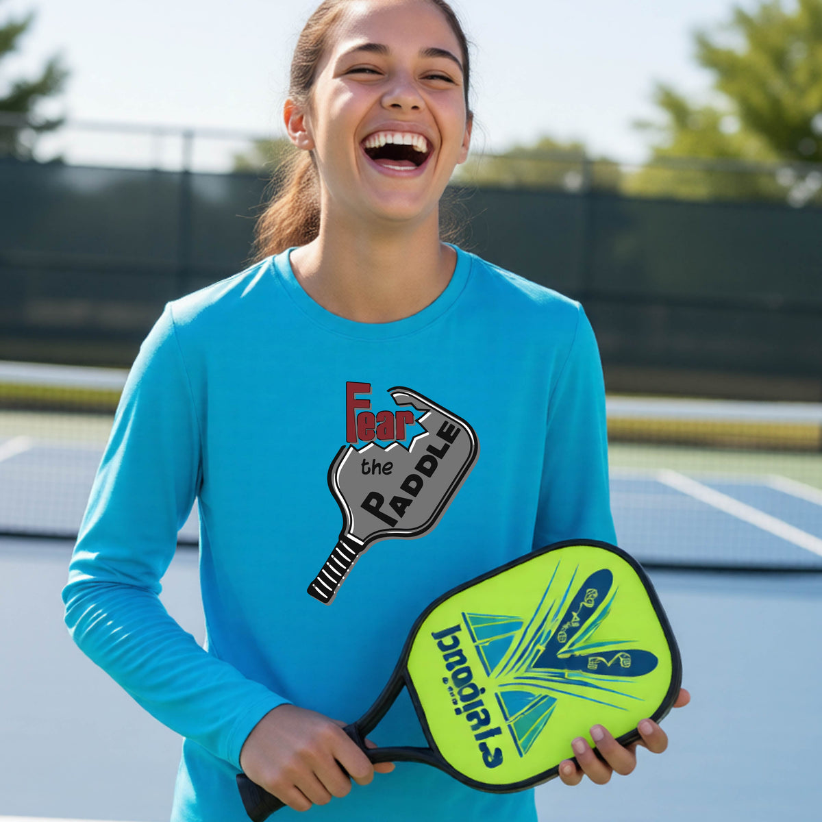 A young person wearing a bright blue long-sleeve shirt with a pickleball graphic of the text 'FEAR THE PADDLE' cut out into a pickleball paddle. 