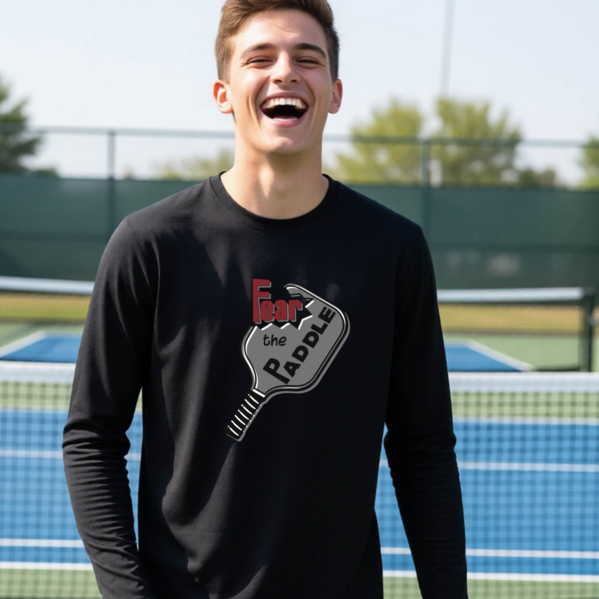 A young person wearing a black long-sleeve shirt with a pickleball graphic of the text 'FEAR THE PADDLE' cut out into a pickleball paddle. 