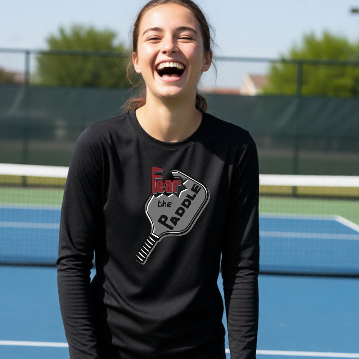 A young person wearing a black long-sleeve shirt with a pickleball graphic of the text 'FEAR THE PADDLE' cut out into a pickleball paddle. 