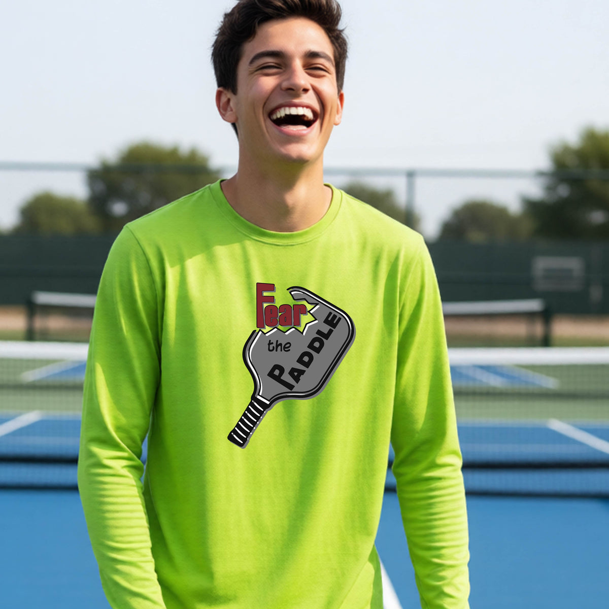 A young person wearing a lime green long-sleeve shirt with a pickleball graphic of the text 'FEAR THE PADDLE' cut out into a pickleball paddle. 