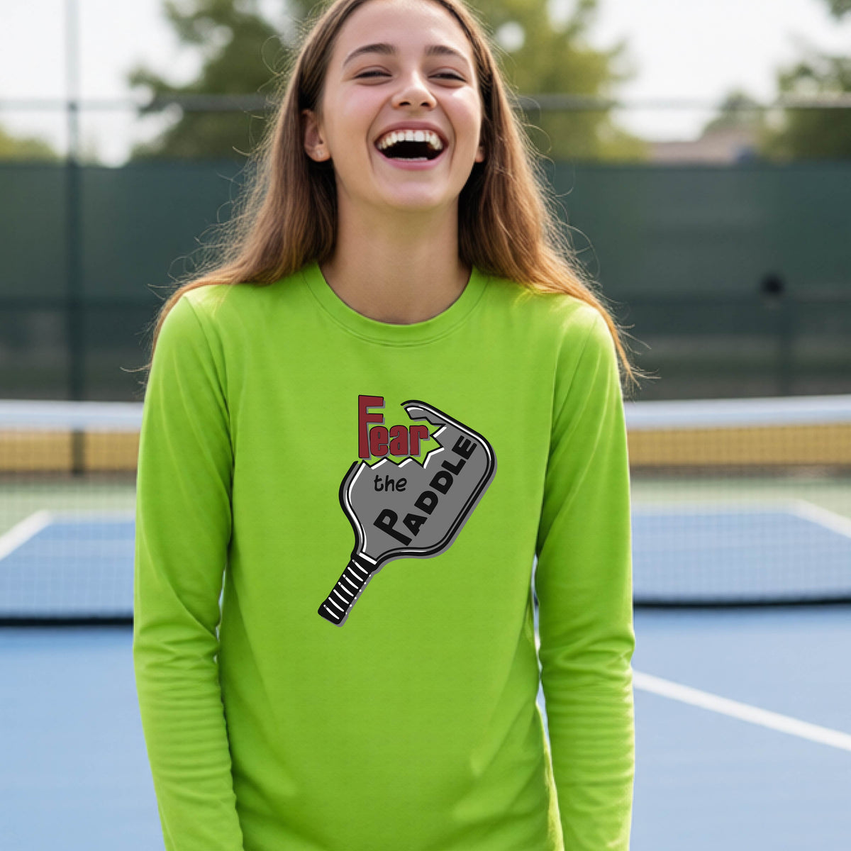 A young person wearing a lime green long-sleeve shirt with a pickleball graphic of the text 'FEAR THE PADDLE' cut out into a pickleball paddle. 