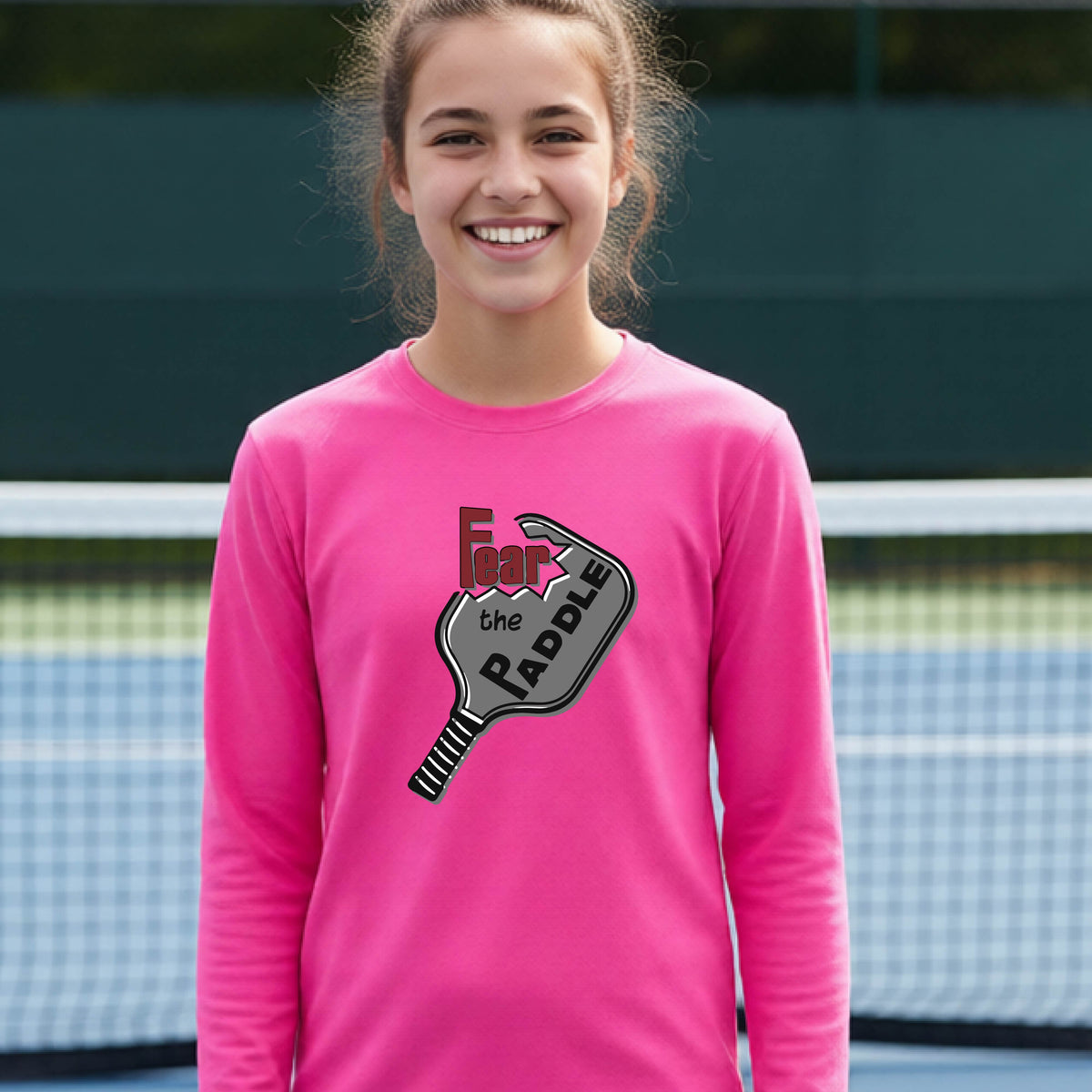 A young person wearing a pink long-sleeve shirt with a pickleball graphic of the text 'FEAR THE PADDLE' cut out into a pickleball paddle. 