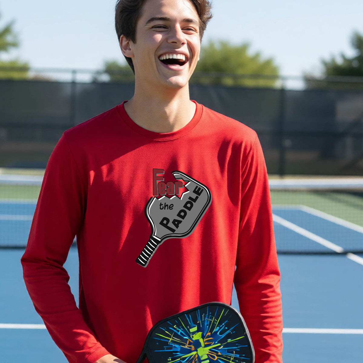 A young person wearing a red long-sleeve shirt with a pickleball graphic of the text 'FEAR THE PADDLE' cut out into a pickleball paddle. 