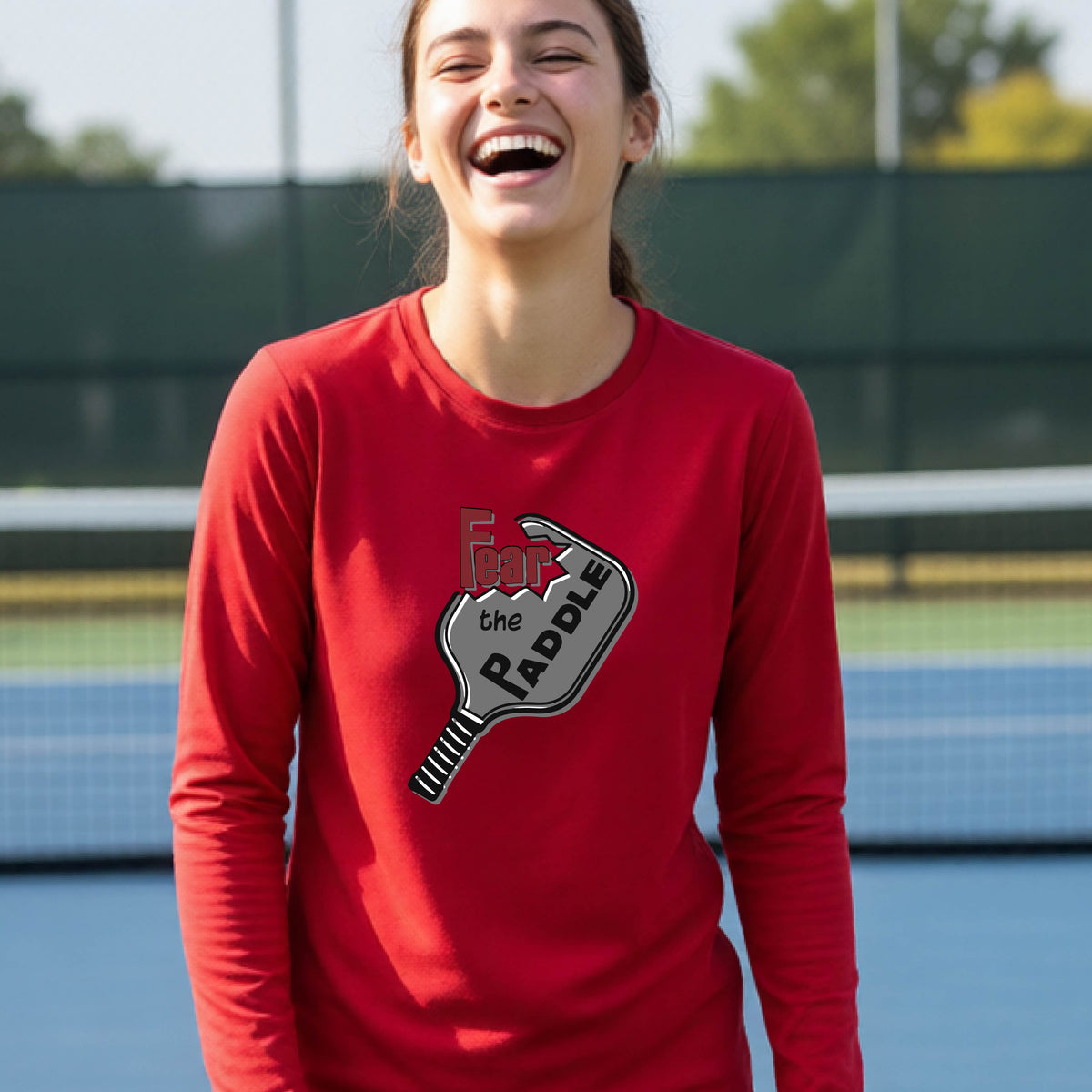 A young person wearing a red long-sleeve shirt with a pickleball graphic of the text 'FEAR THE PADDLE' cut out into a pickleball paddle. 