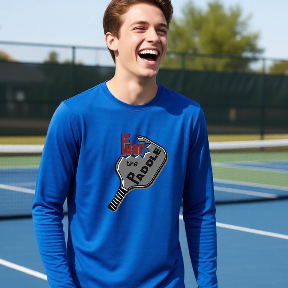 A young person wearing a royal blue long-sleeve shirt with a pickleball graphic of the text 'FEAR THE PADDLE' cut out into a pickleball paddle. 