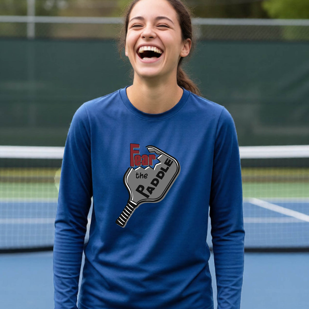 A young person wearing a royal blue long-sleeve shirt with a pickleball graphic of the text 'FEAR THE PADDLE' cut out into a pickleball paddle. 