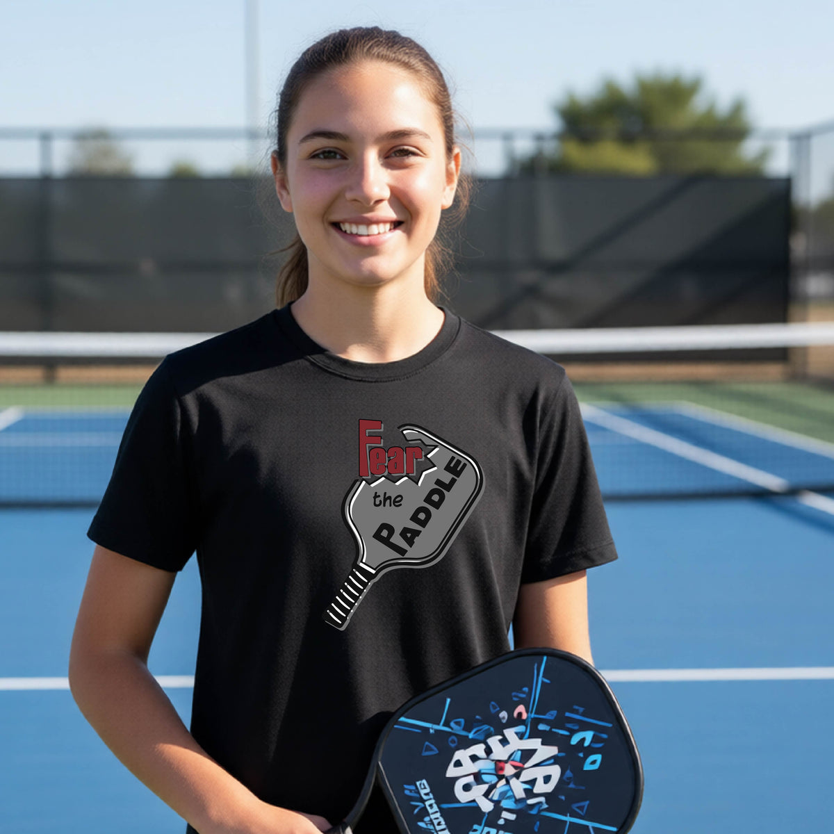 A young person smiling on a blue pickleball court, wearing a black t-shirt with a graphic that reads 'Fear the PADDLE' over a pickleball paddle design.