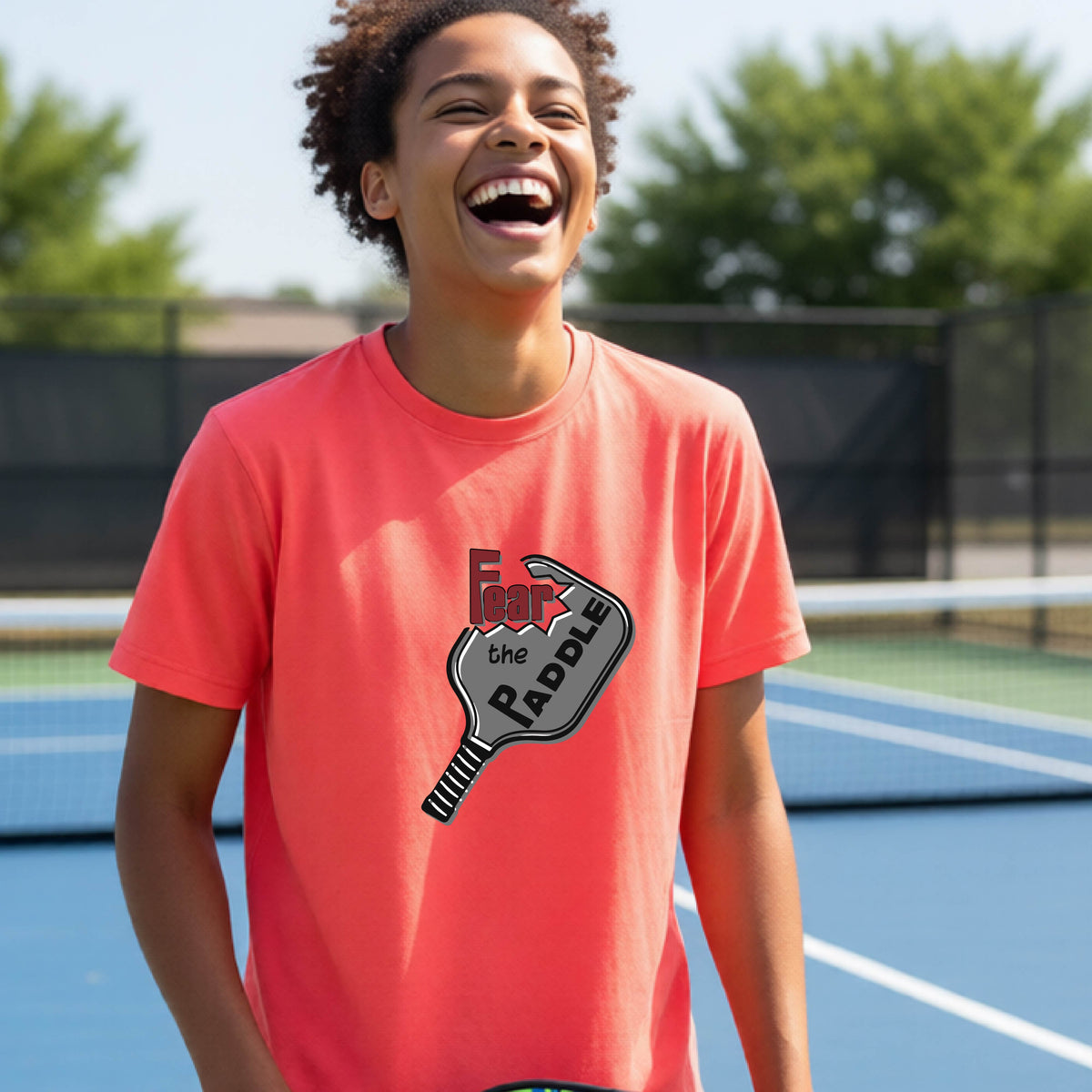 A young person smiling on a blue pickleball court, wearing a coral pink t-shirt with a graphic that reads 'Fear the PADDLE' over a pickleball paddle design.
