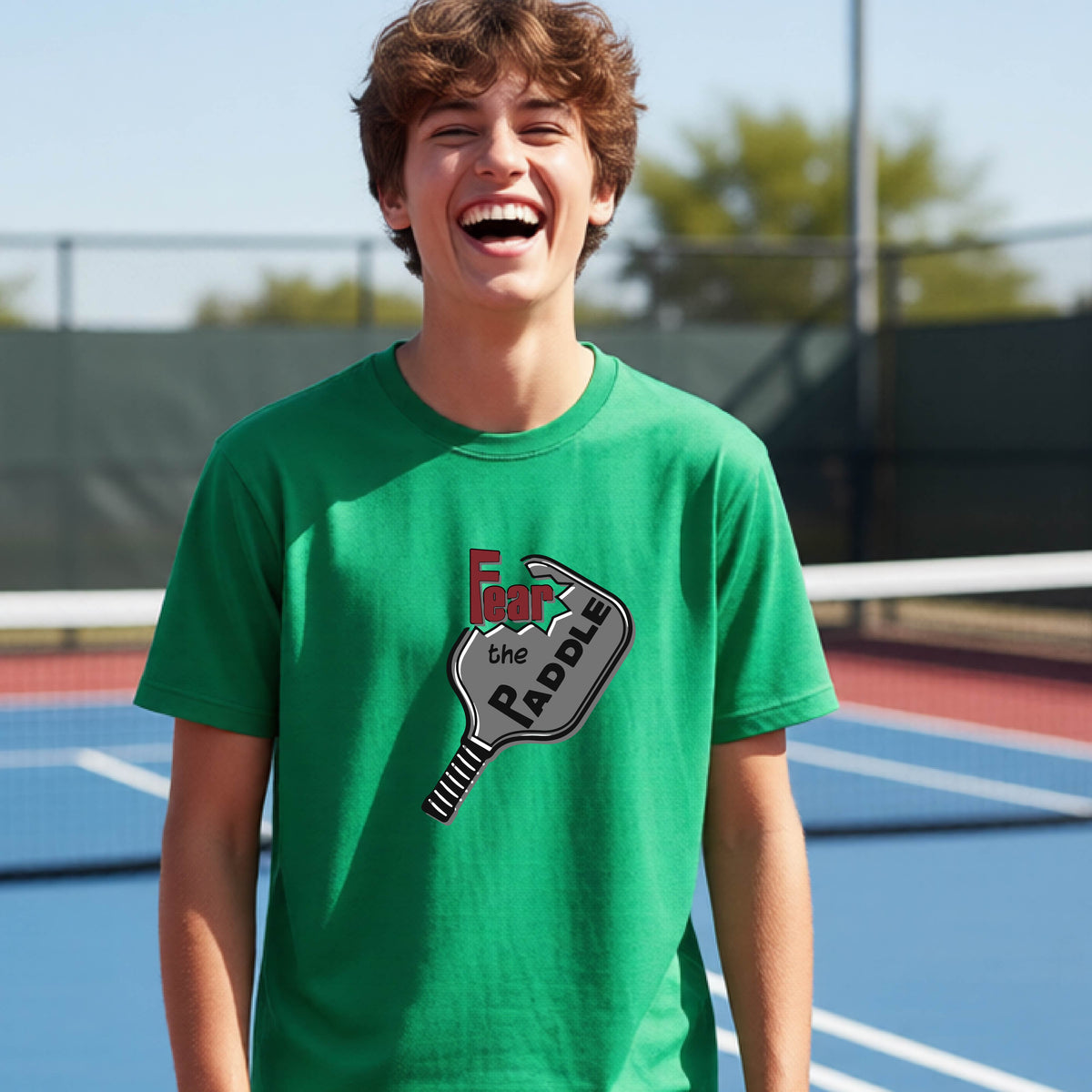 A young person smiling on a blue pickleball court, wearing a green t-shirt with a graphic that reads 'Fear the PADDLE' over a pickleball paddle design.