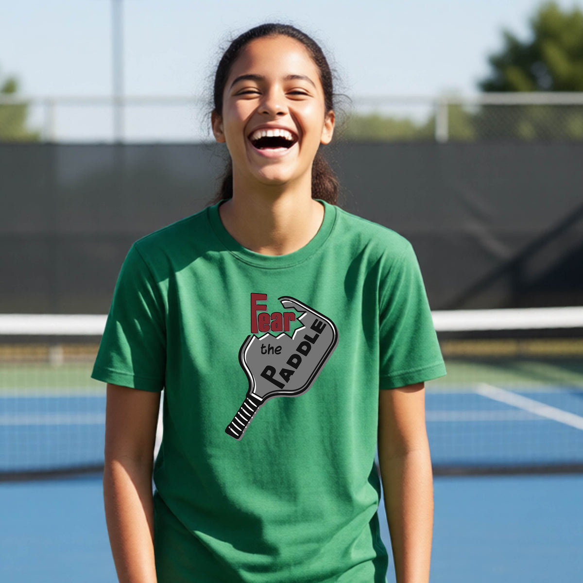 A young person smiling on a blue pickleball court, wearing a green t-shirt with a graphic that reads 'Fear the PADDLE' over a pickleball paddle design.