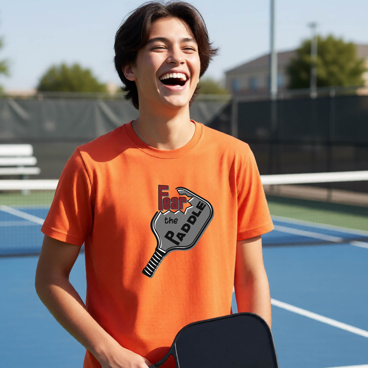 A young person smiling on a blue pickleball court, wearing an orange t-shirt with a graphic that reads 'Fear the PADDLE' over a pickleball paddle design.