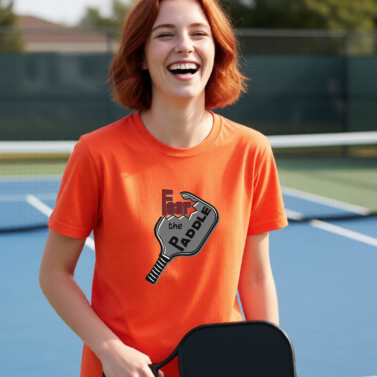 A young person smiling on a blue pickleball court, wearing a bright orange t-shirt with a graphic that reads 'Fear the PADDLE' over a pickleball paddle design.