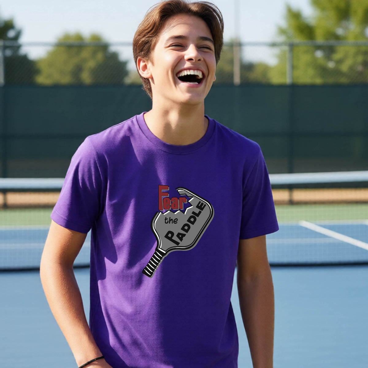A young person smiling on a blue pickleball court, wearing a purple t-shirt with a graphic that reads 'Fear the PADDLE' over a pickleball paddle design.