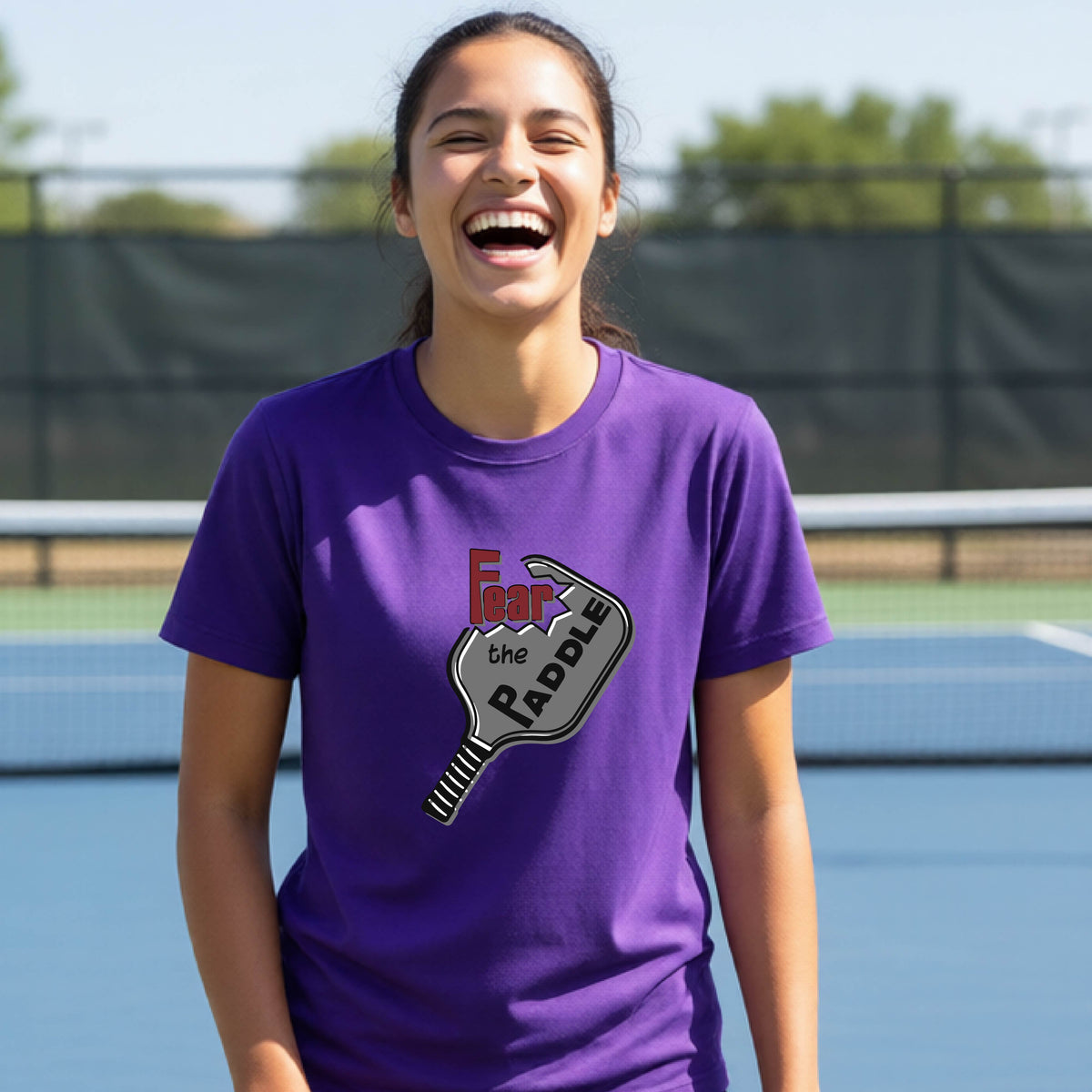 A young person smiling on a blue pickleball court, wearing a purple t-shirt with a graphic that reads 'Fear the PADDLE' over a pickleball paddle design.