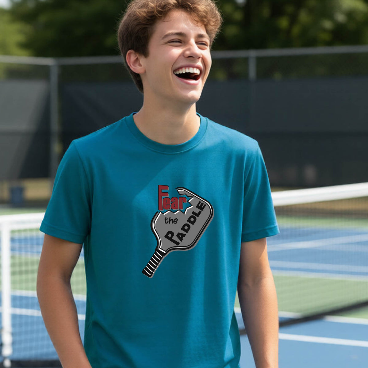 A young person smiling on a blue pickleball court, wearing a teal t-shirt with a graphic that reads 'Fear the PADDLE' over a pickleball paddle design.