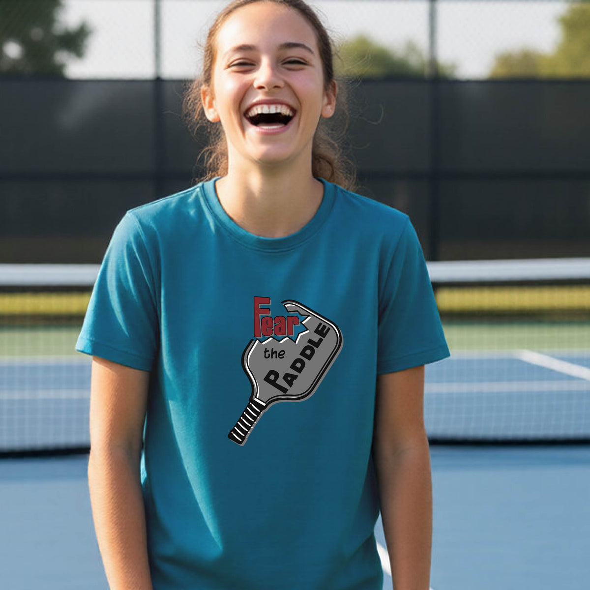 A young person smiling on a blue pickleball court, wearing a teal t-shirt with a graphic that reads 'Fear the PADDLE' over a pickleball paddle design.