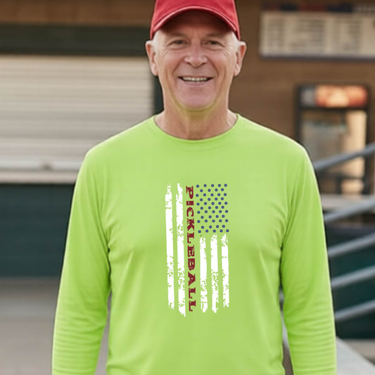 A man wearing a lime green short-sleeve t-shirt with a pickleball graphic of a distressed flag made out of pickleballs and the text 'PICKLEBALL' replacing one of the stripes, standing on a pickleball court.