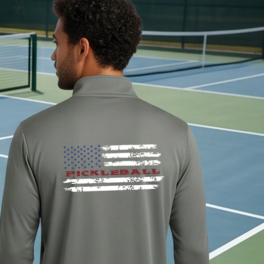 A man wearing a gray quarter-zip shirt with a pickleball graphic on the back of a distressed flag made out of pickleballs and the text 'PICKLEBALL' replacing one of the stripes, standing on a pickleball court.