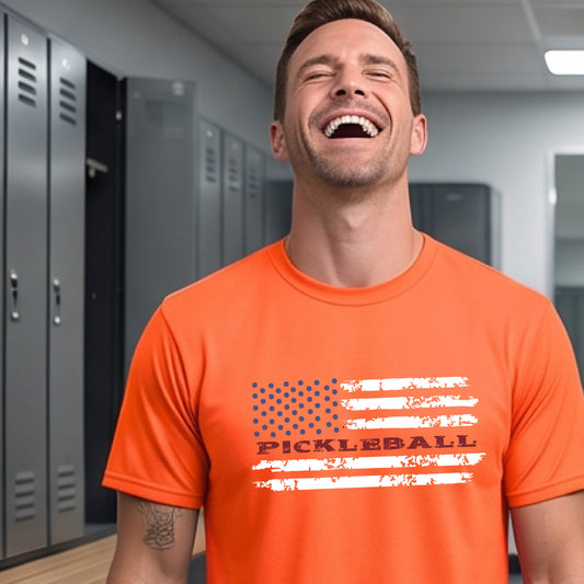 A man wearing an orange short-sleeve t-shirt with a pickleball graphic of a distressed flag made out of pickleballs and the text 'PICKLEBALL' replacing one of the stripes, standing on a pickleball court.