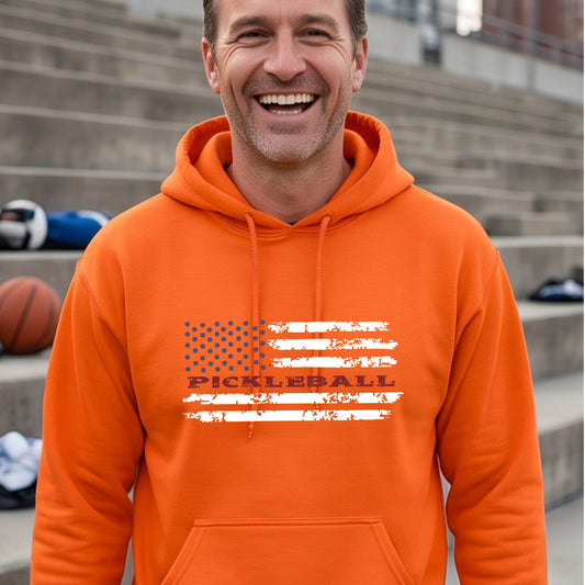 A man wearing a unisex orange hoodie pullover with a pickleball graphic of a distressed flag made out of pickleballs and the text 'PICKLEBALL' replacing one of the stripes, standing on a pickleball court.