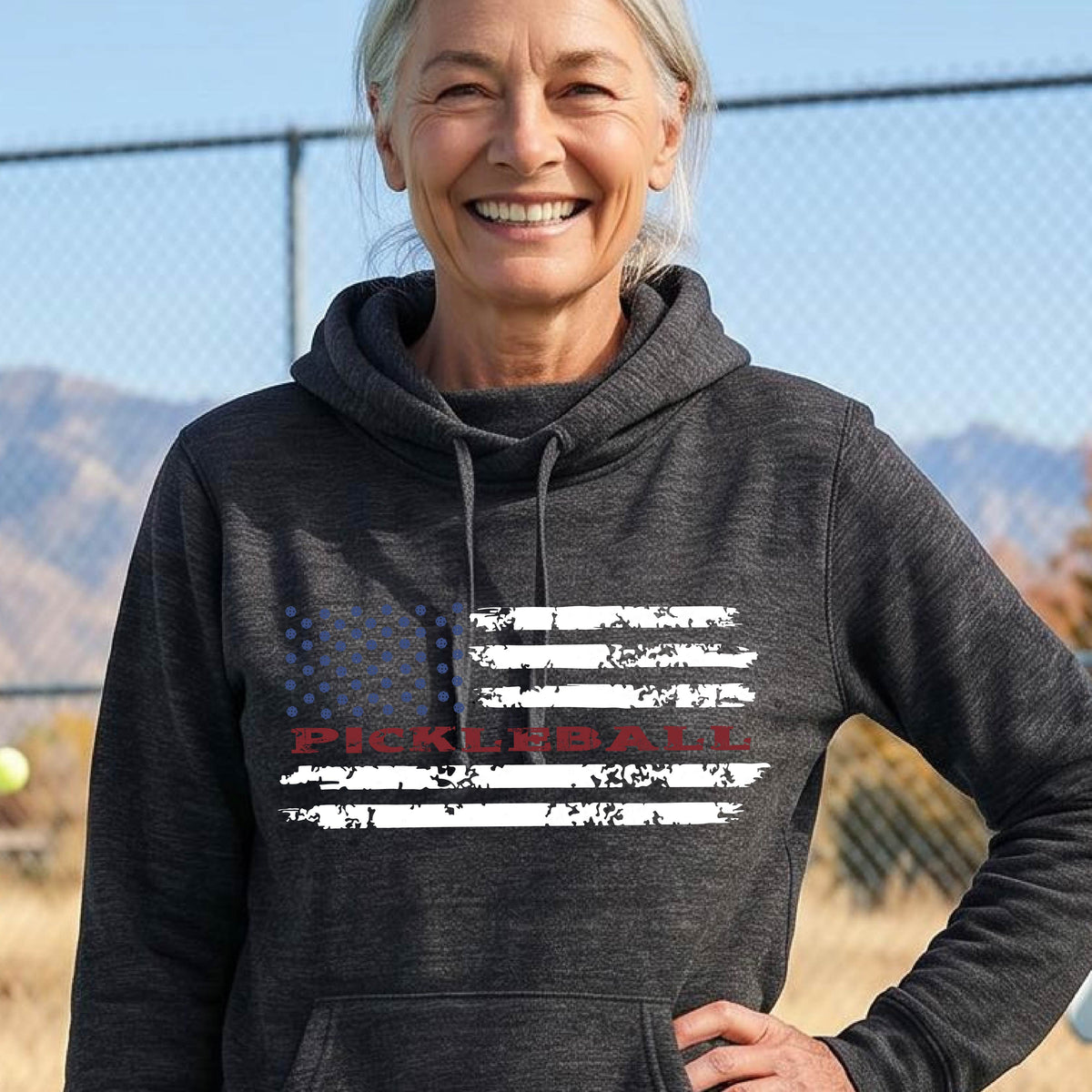A woman wearing a black cowl-neck pullover hoodies with a pickleball graphic of a distressed flag made out of pickleballs and the text 'PICKLEBALL' replacing one of the stripes, standing on a pickleball court.