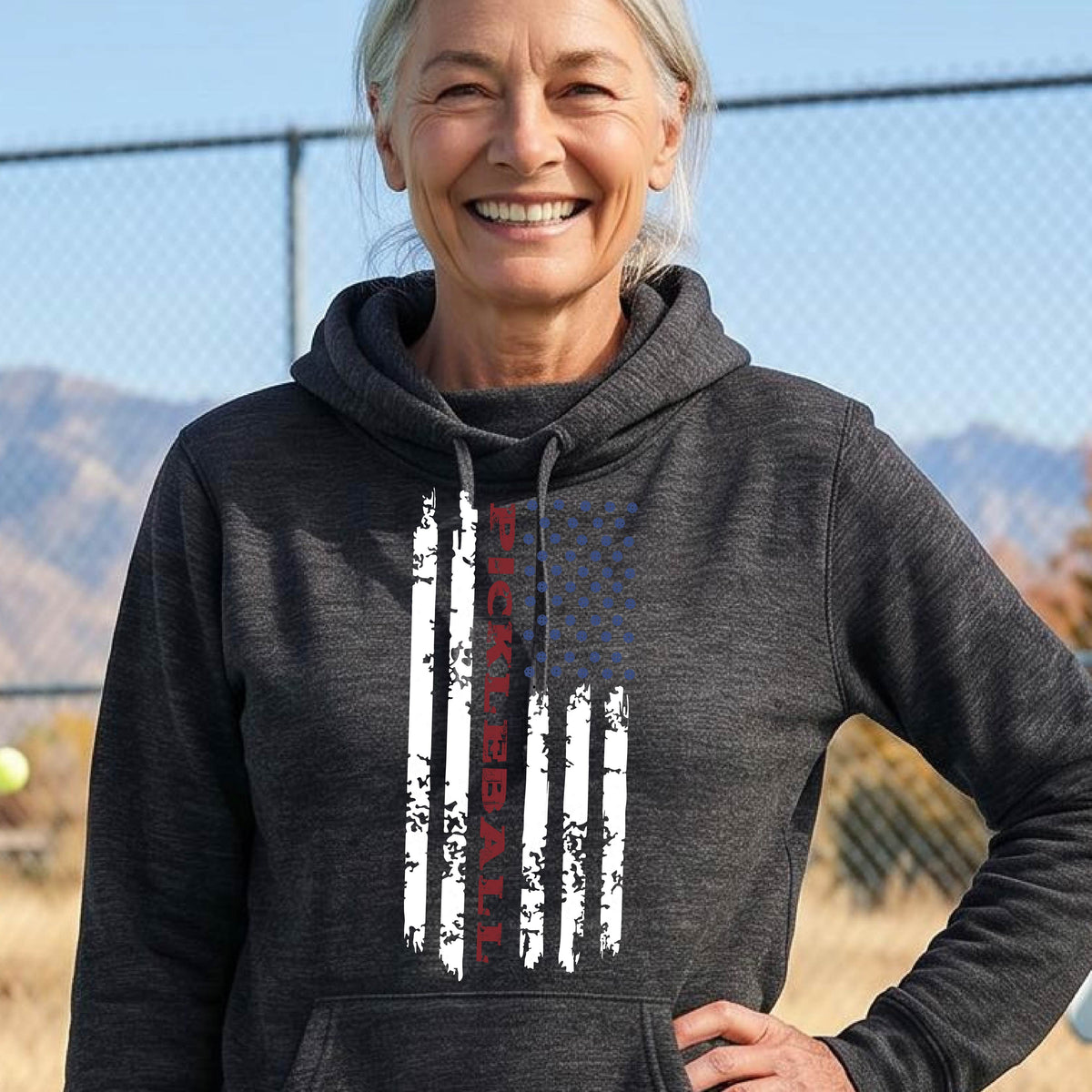 A woman wearing a black cowl-neck pullover hoodies with a pickleball graphic of a distressed flag made out of pickleballs and the text 'PICKLEBALL' replacing one of the stripes, standing on a pickleball court.