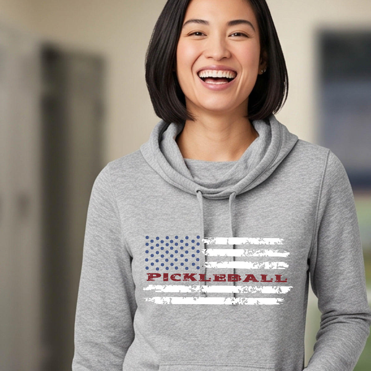 A woman wearing a gray cowl-neck pullover hoodies with a pickleball graphic of a distressed flag made out of pickleballs and the text 'PICKLEBALL' replacing one of the stripes, standing on a pickleball court.