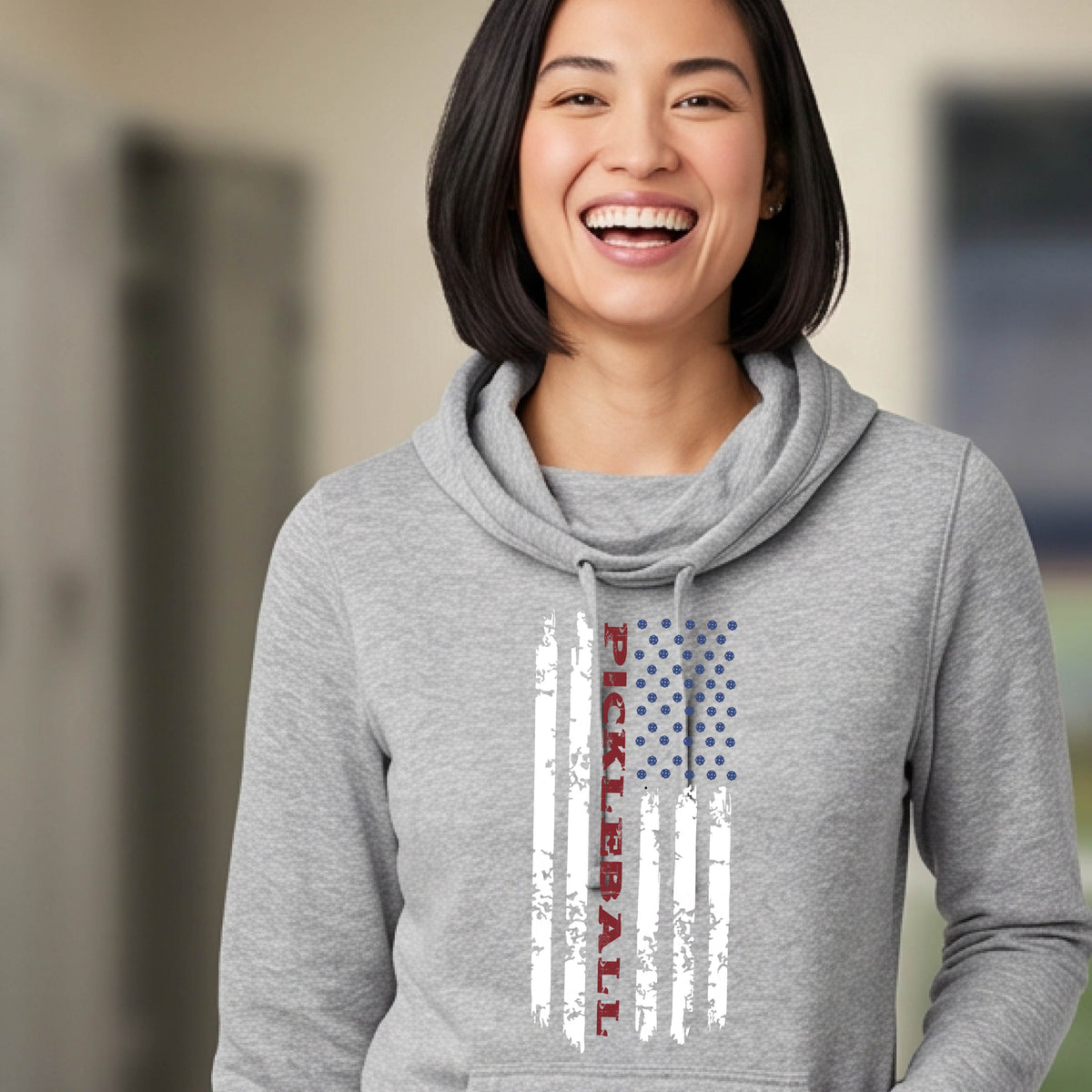 A woman wearing a gray cowl-neck pullover hoodies with a pickleball graphic of a distressed flag made out of pickleballs and the text 'PICKLEBALL' replacing one of the stripes, standing on a pickleball court.