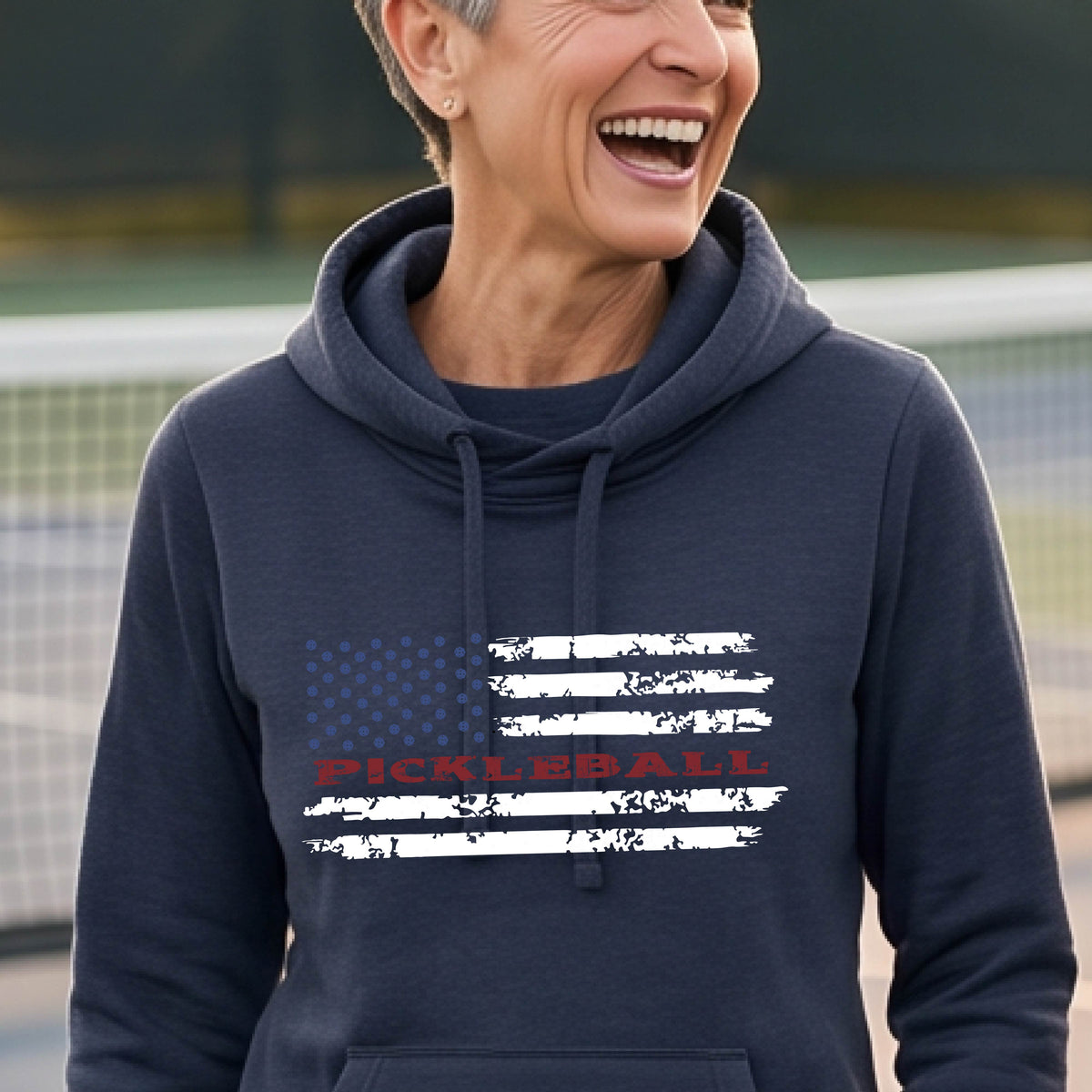 A woman wearing a navy blue cowl-neck pullover hoodies with a pickleball graphic of a distressed flag made out of pickleballs and the text 'PICKLEBALL' replacing one of the stripes, standing on a pickleball court.