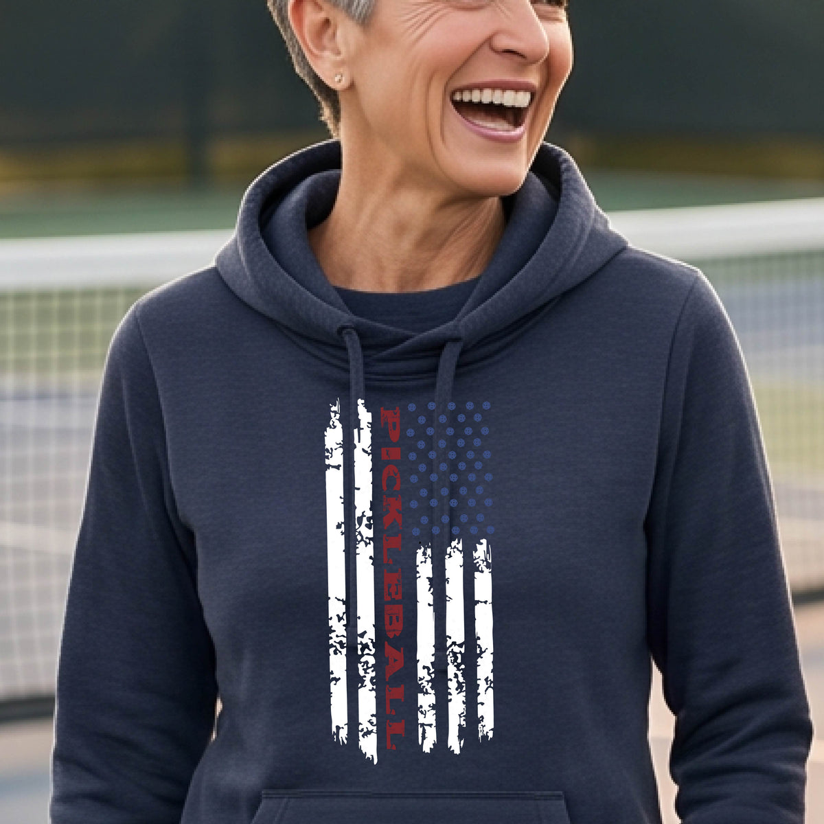 A woman wearing a navy blue cowl-neck pullover hoodies with a pickleball graphic of a distressed flag made out of pickleballs and the text 'PICKLEBALL' replacing one of the stripes, standing on a pickleball court.