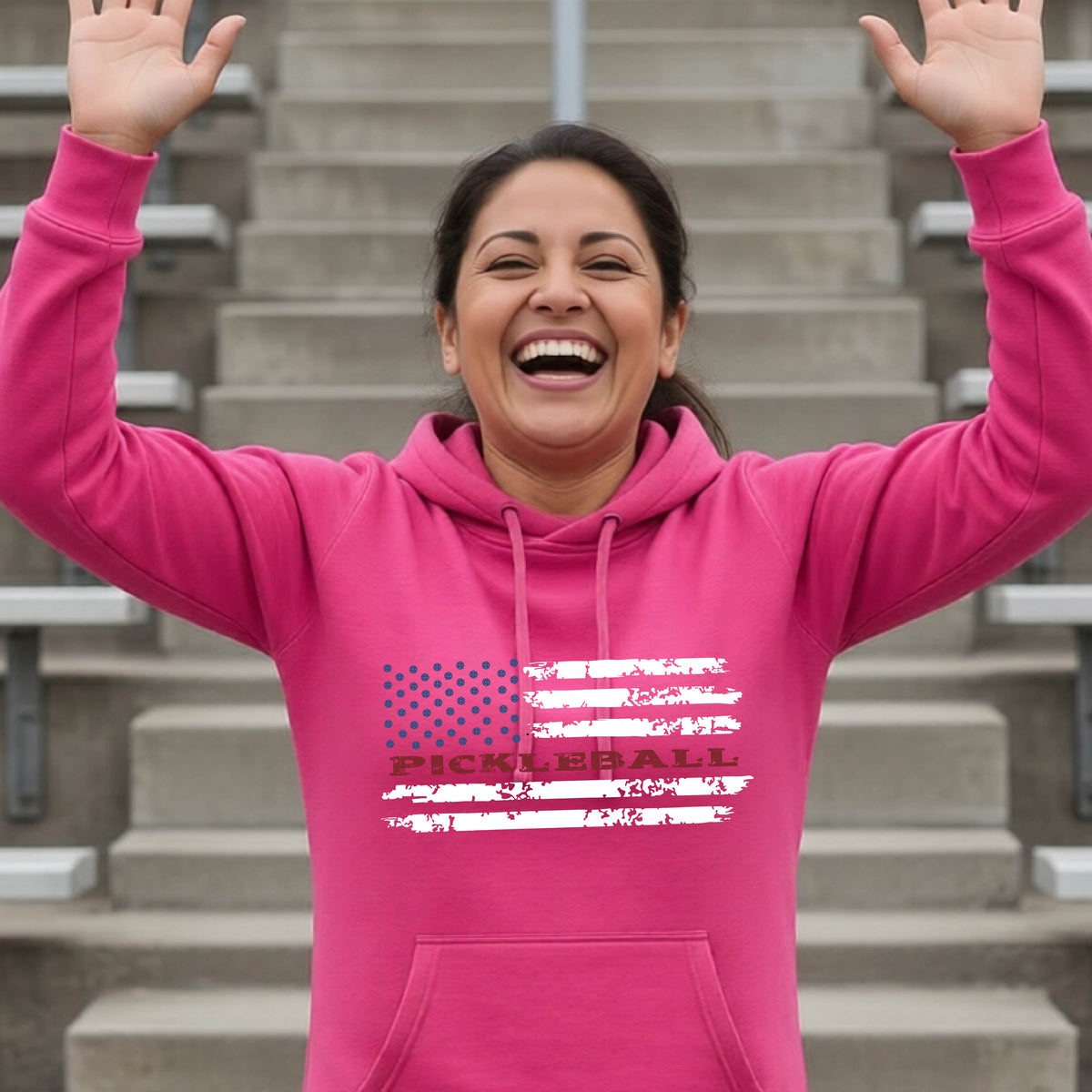 A woman wearing a pink cowl-neck pullover hoodies with a pickleball graphic of a distressed flag made out of pickleballs and the text 'PICKLEBALL' replacing one of the stripes, standing on a pickleball court.