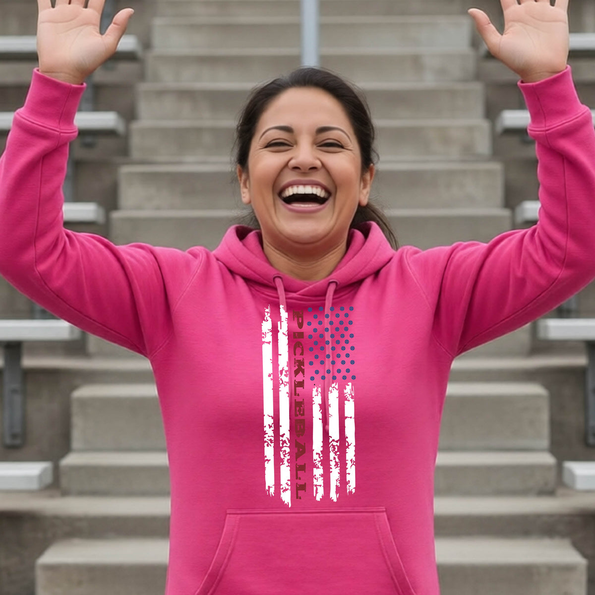 A woman wearing a pink cowl-neck pullover hoodies with a pickleball graphic of a distressed flag made out of pickleballs and the text 'PICKLEBALL' replacing one of the stripes, standing on a pickleball court.