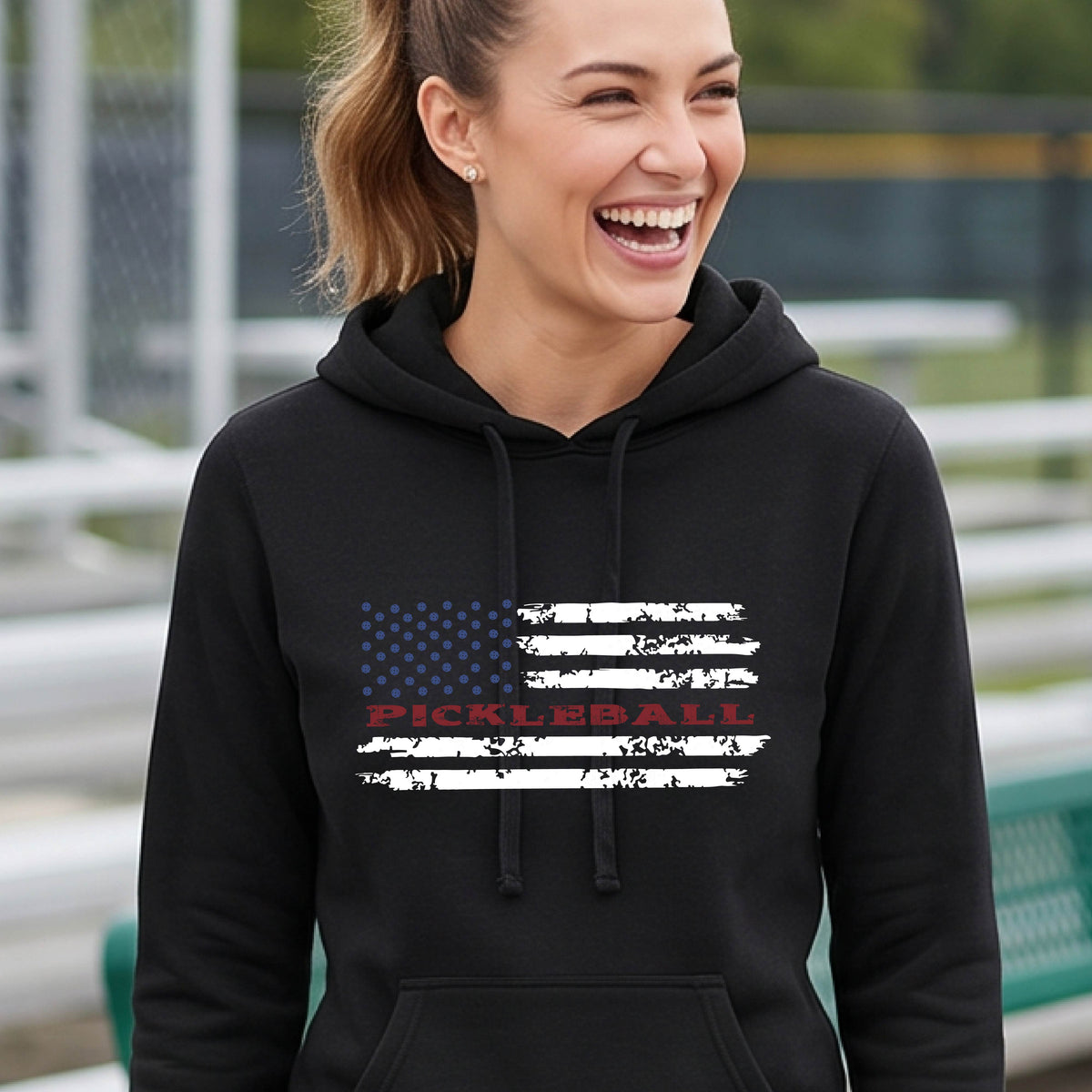 A woman wearing a black fitted hoodie pullover with a pickleball graphic of a distressed flag made out of pickleballs and the text 'PICKLEBALL' replacing one of the stripes, standing on a pickleball court.