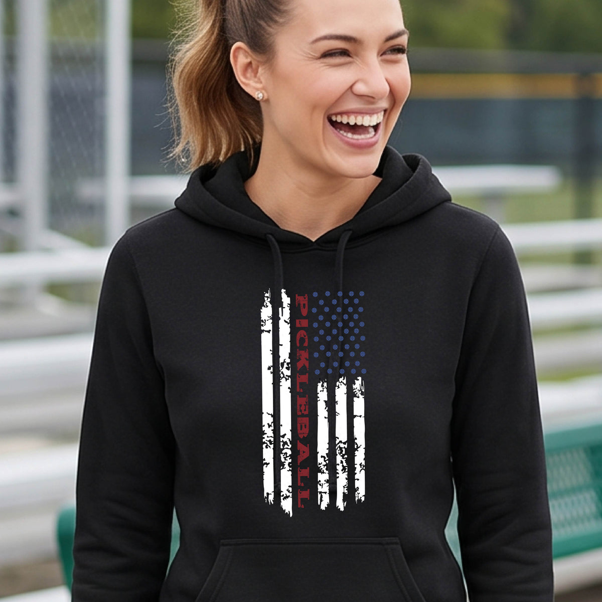 A woman wearing a black fitted hoodie pullover with a pickleball graphic of a distressed flag made out of pickleballs and the text 'PICKLEBALL' replacing one of the stripes, standing on a pickleball court.