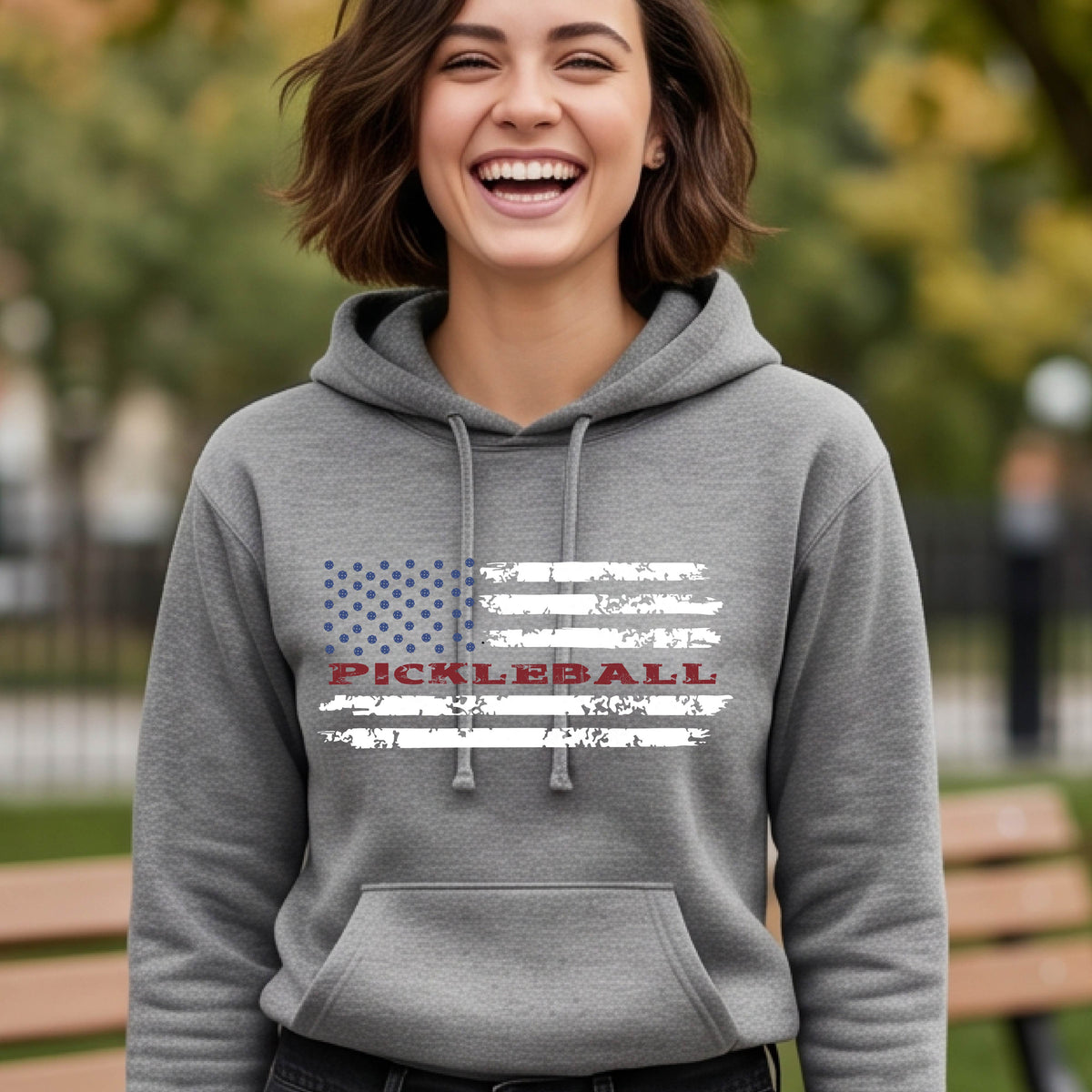 A woman wearing a gray fitted hoodie pullover with a pickleball graphic of a distressed flag made out of pickleballs and the text 'PICKLEBALL' replacing one of the stripes, standing on a pickleball court.