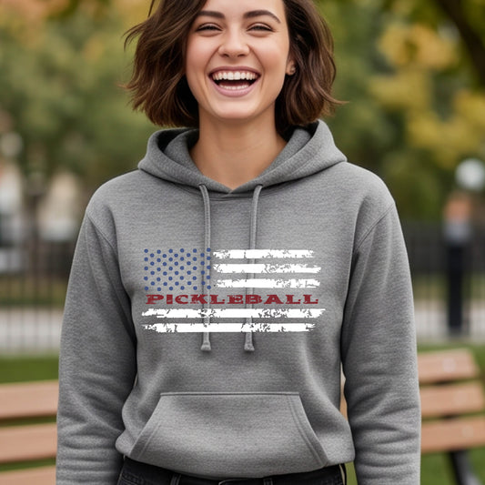 A woman wearing a gray fitted hoodie pullover with a pickleball graphic of a distressed flag made out of pickleballs and the text 'PICKLEBALL' replacing one of the stripes, standing on a pickleball court.