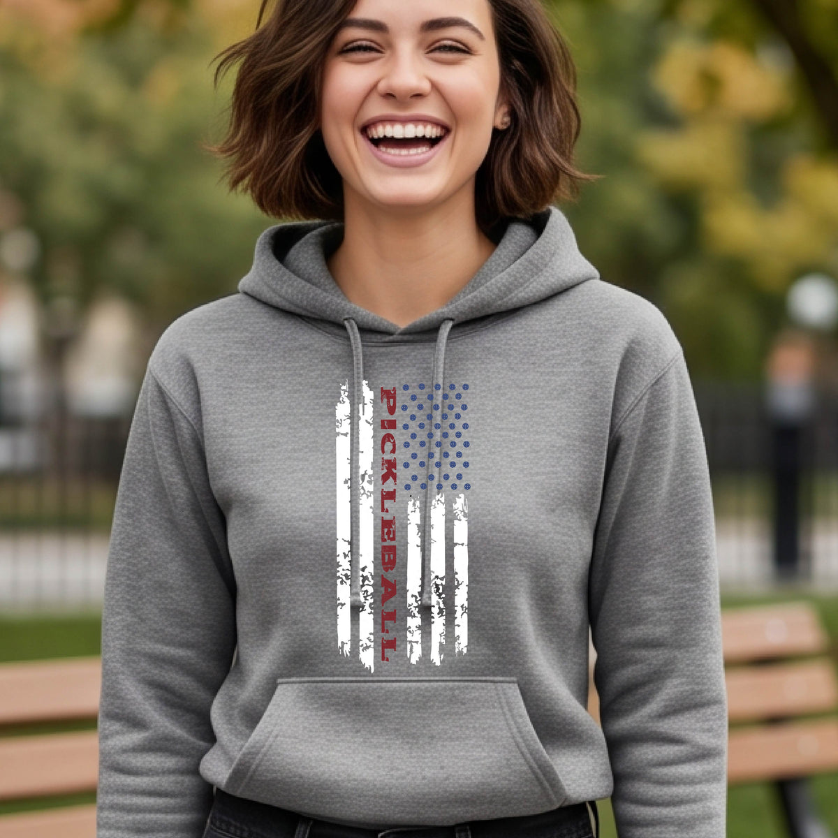 A woman wearing a gray fitted hoodie pullover with a pickleball graphic of a distressed flag made out of pickleballs and the text 'PICKLEBALL' replacing one of the stripes, standing on a pickleball court.