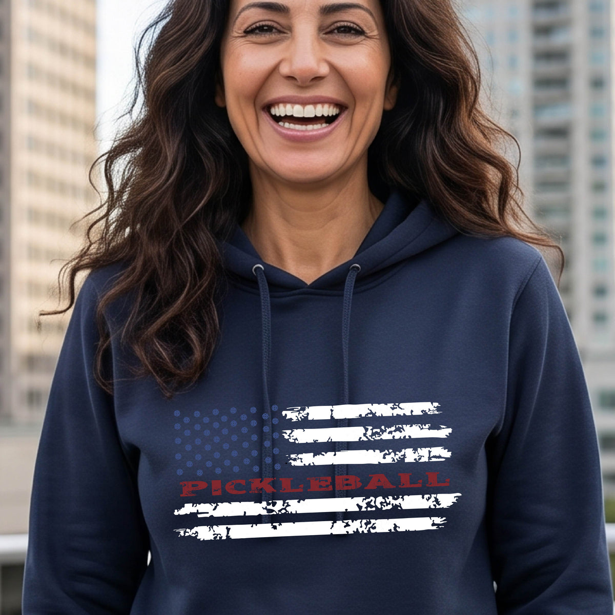 A woman wearing a navy blue fitted hoodie pullover with a pickleball graphic of a distressed flag made out of pickleballs and the text 'PICKLEBALL' replacing one of the stripes, standing on a pickleball court.